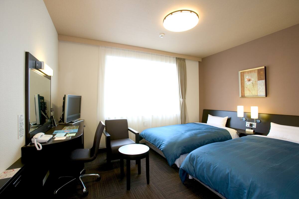 Ein Hotelzimmer mit zwei Betten, einem Schreibtisch mit Telefon, einem Sessel, einem kleinen Tisch, einem Fernseher und einem Bild an der Wand.