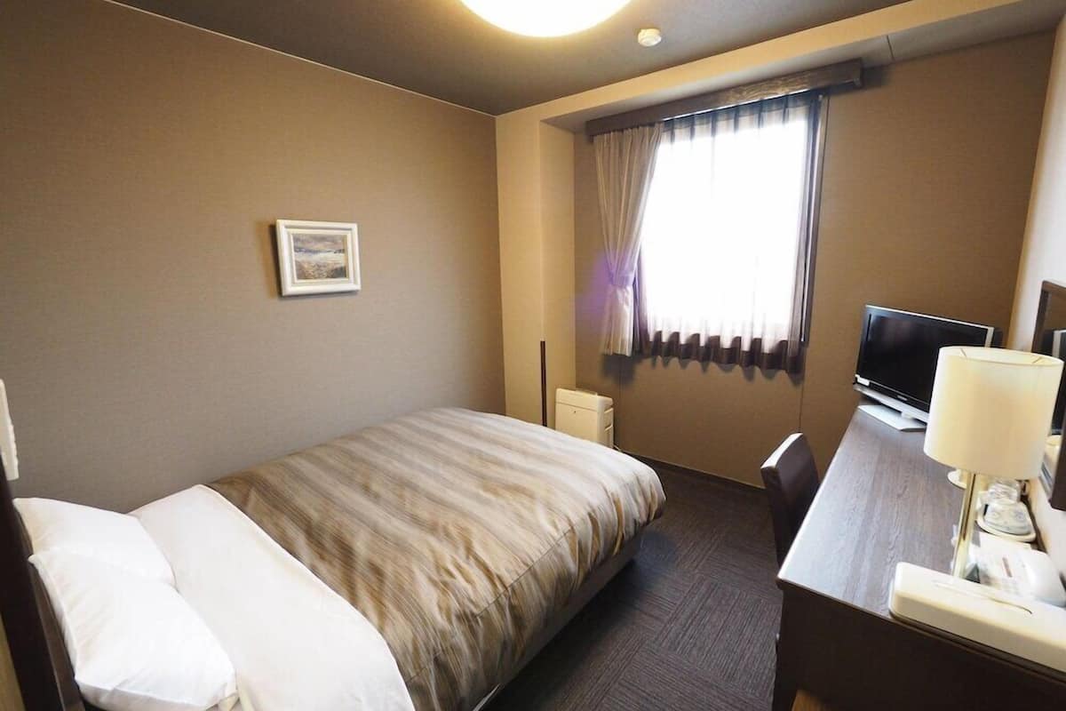 Een hotelkamer met een bed, een bureau met een televisie, een lamp en een raam met gordijnen.