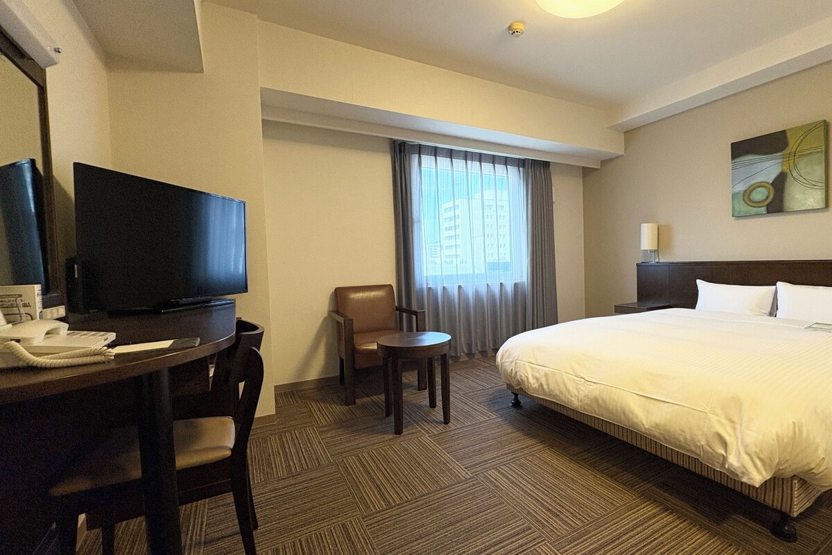 Een hotelkamer met een bed, een bureau met telefoon, een stoel, een televisie en een raam met gordijnen.