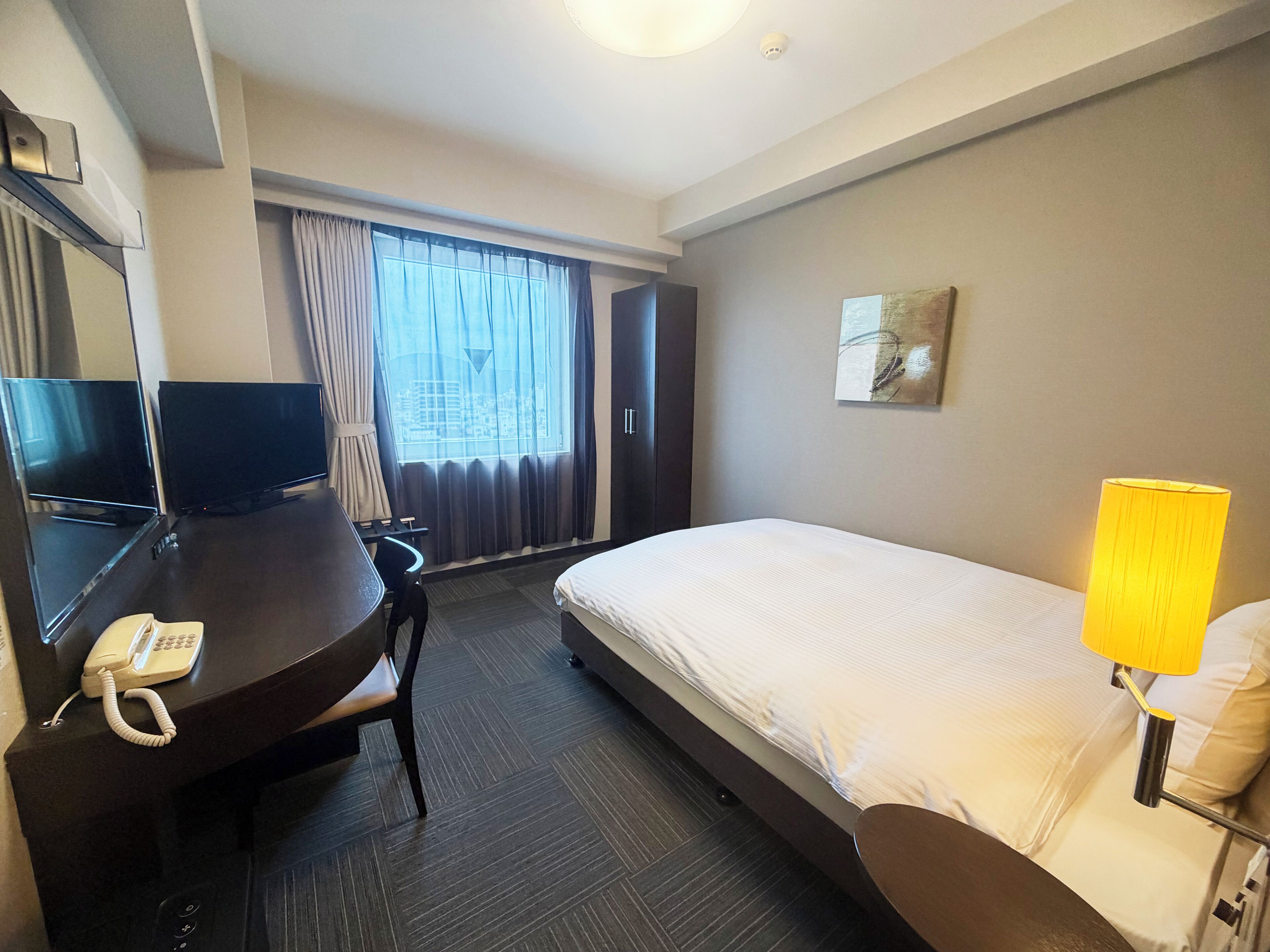 A hotel room with a bed, a desk with a telephone, a chair, a television, and a window with curtains.