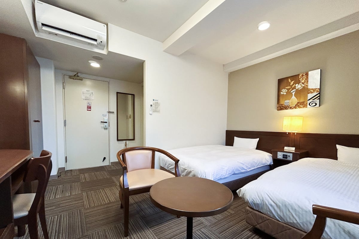 A hotel room with two beds, a desk, a chair, a small table, a lamp, and an air conditioning unit.