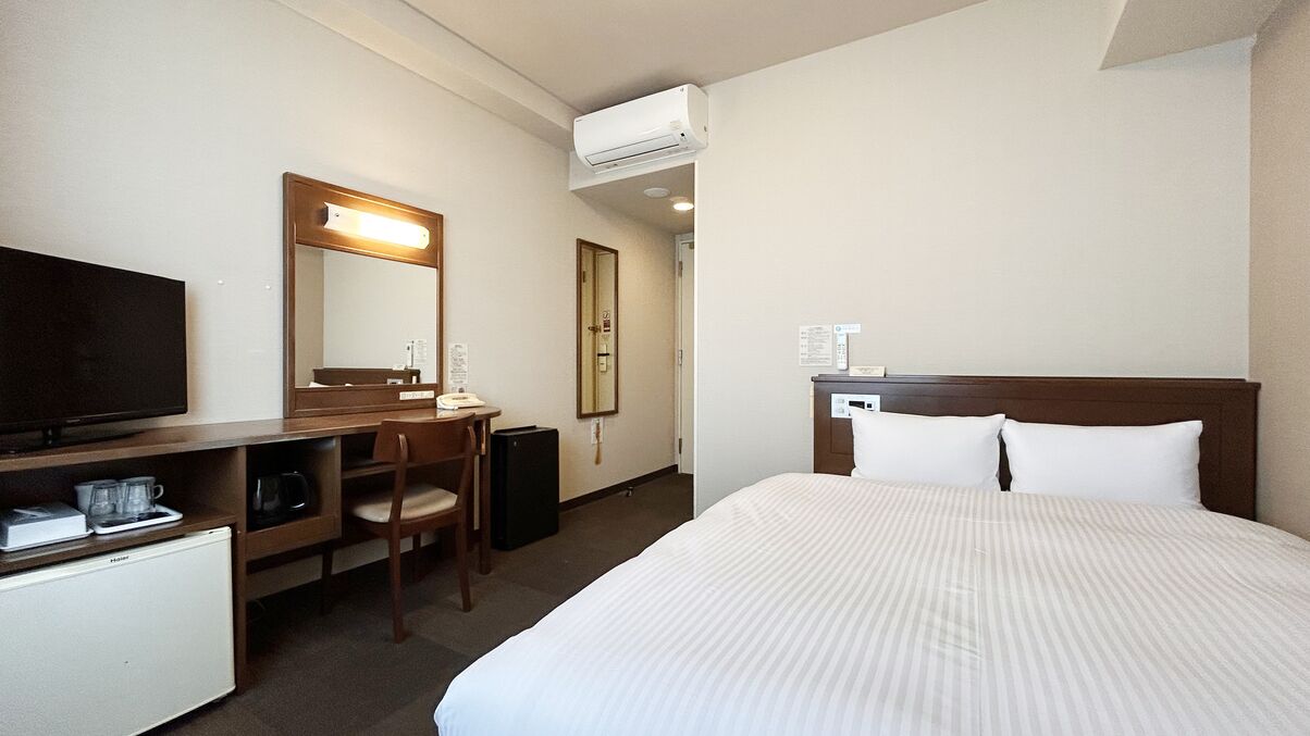 A hotel room with a bed, a desk with a chair, a television, a mini-fridge, and an air conditioning unit.