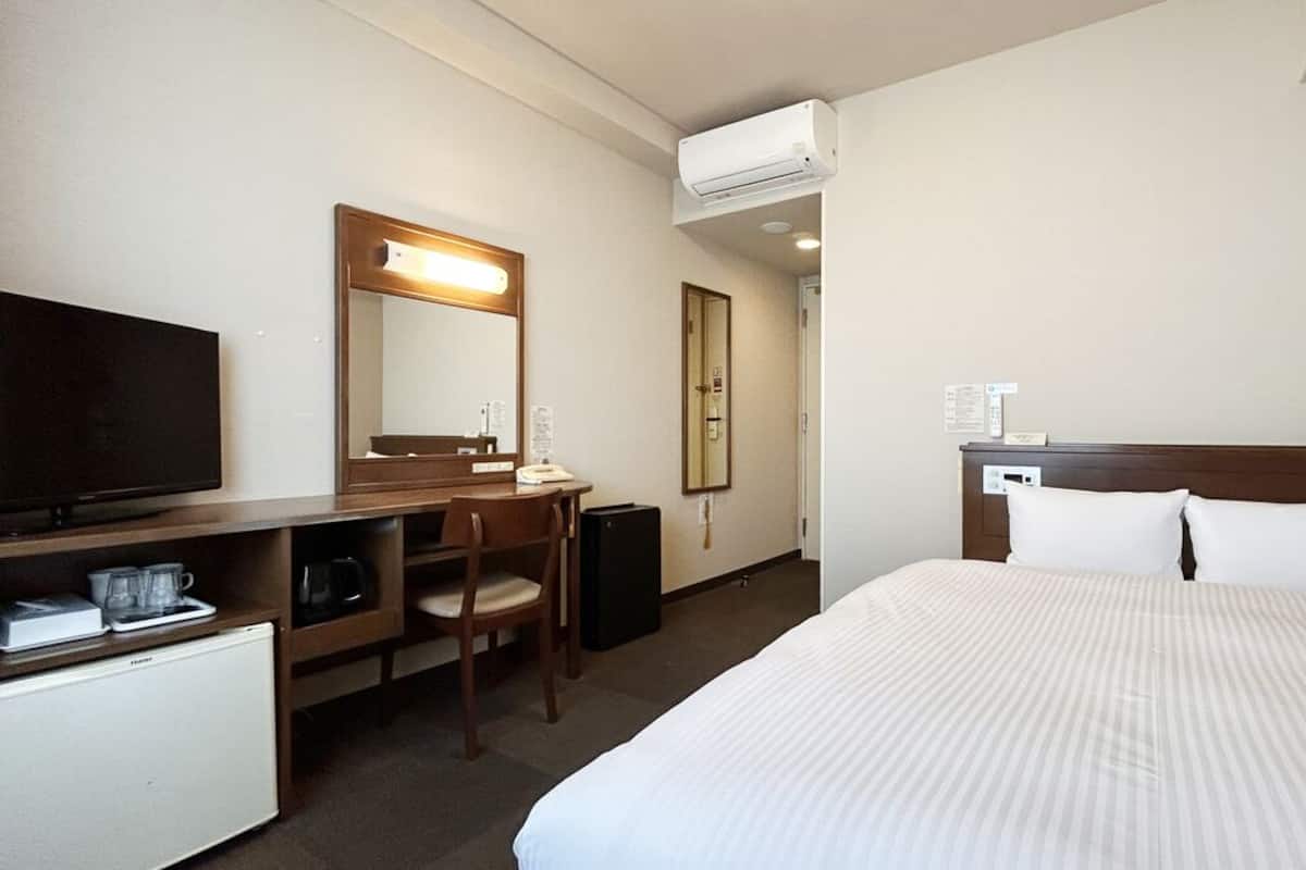 A hotel room with a bed, a desk with a chair, a television, a mini-fridge, and an air conditioning unit.