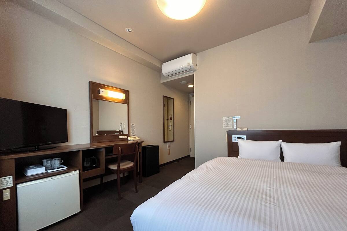 A hotel room with a bed, a desk with a television, a chair, a small refrigerator, and an air conditioning unit.