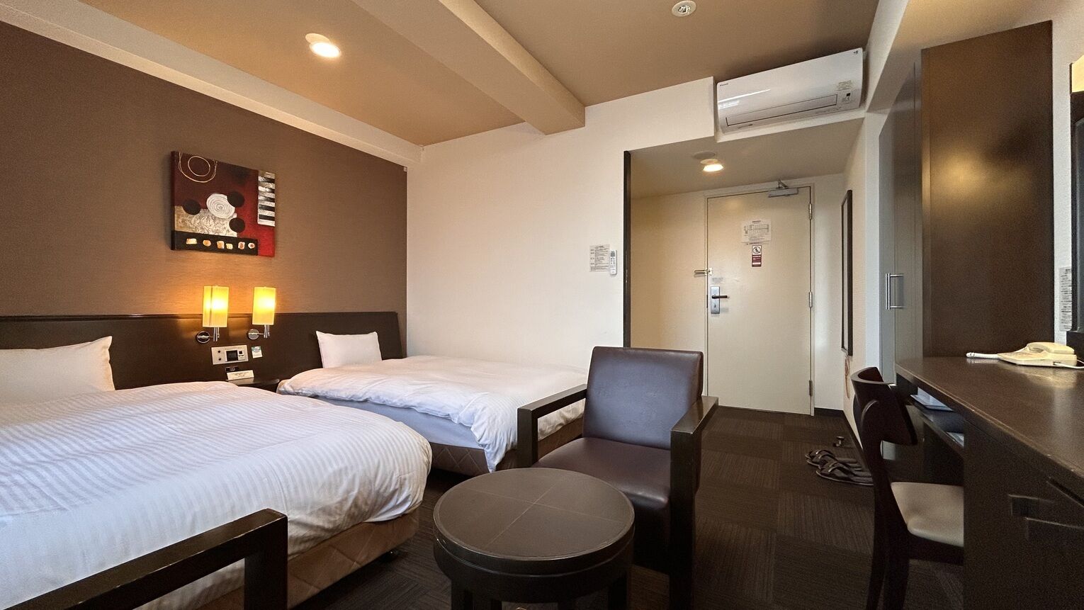 A hotel room with two beds, a desk, a chair, and a wall-mounted artwork.
