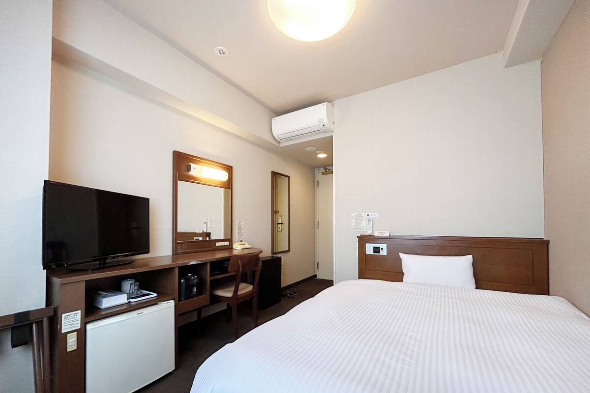A hotel room with a bed, a desk with a television, a mirror, and a small refrigerator.
