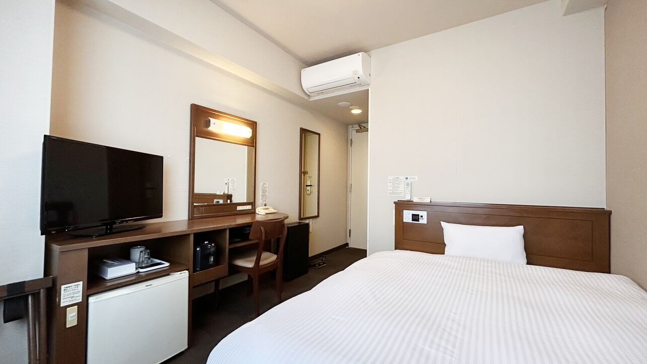 A hotel room with a bed, a desk with a computer, a television, a mini-fridge, and a mirror.
