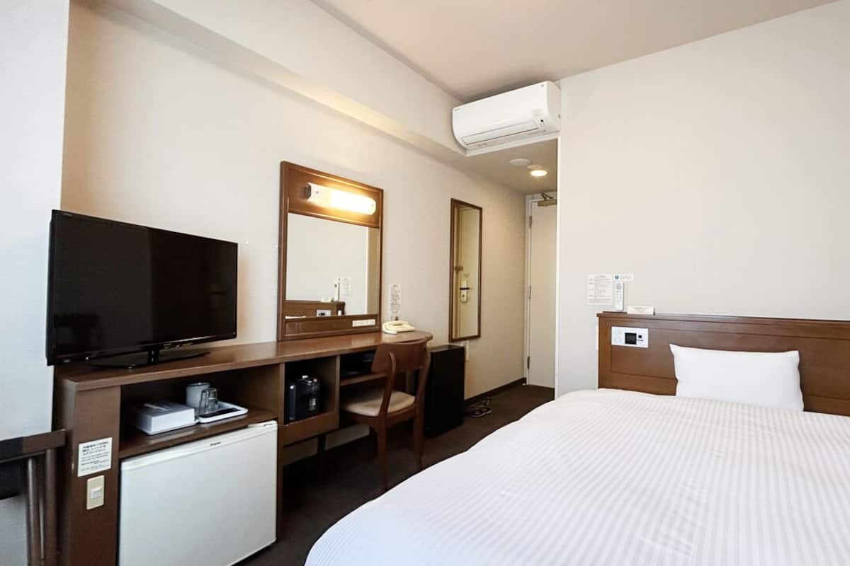 A hotel room with a bed, a desk with a computer, a television, a mini-fridge, and a mirror.