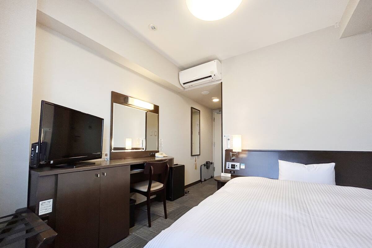 A hotel room with a bed, a desk with a chair, a television, a mirror, an air conditioning unit, and a bedside table with a lamp.