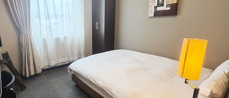 A hotel room with a bed, a desk, a chair, a wardrobe, and a wall-mounted lamp.