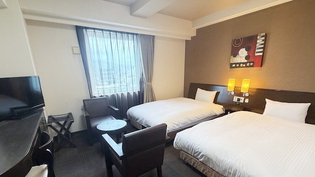 Habitación de hotel con dos camas, un escritorio, una silla, una mesita, un televisor y una ventana con cortinas.