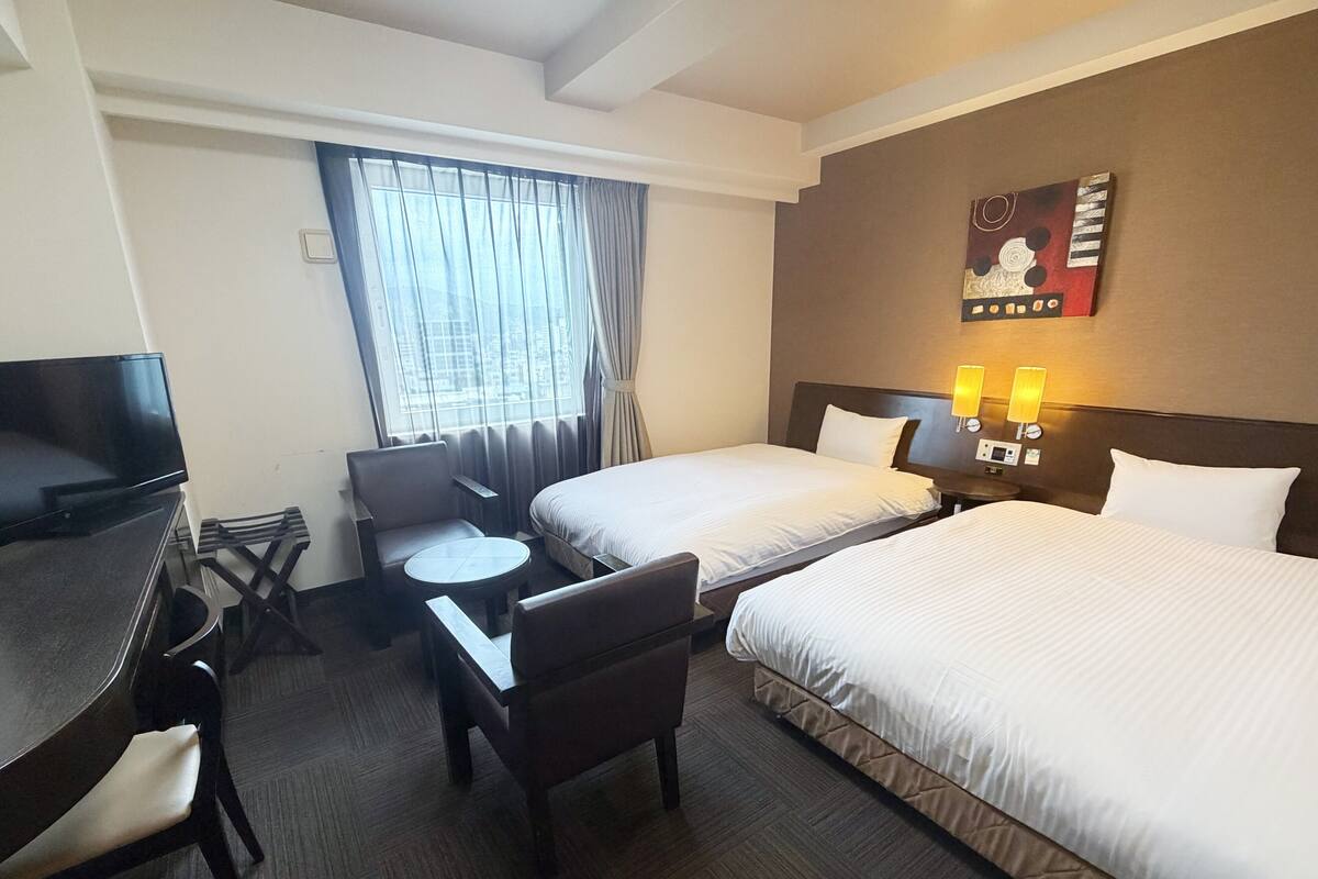 A hotel room with two beds, a desk, a chair, a small table, a TV, and a window with curtains.