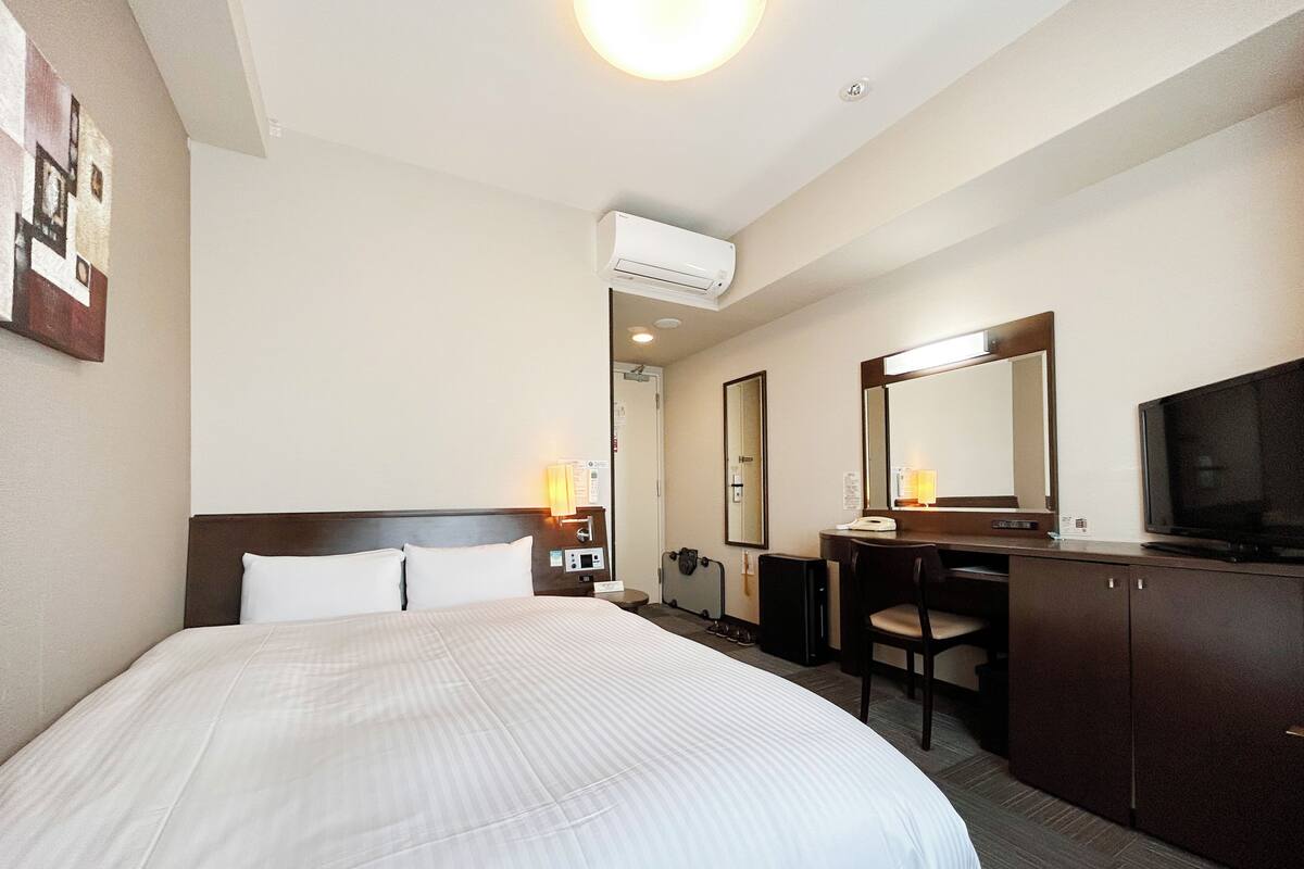 A hotel room with a bed, a desk, a chair, a television, and an air conditioning unit.