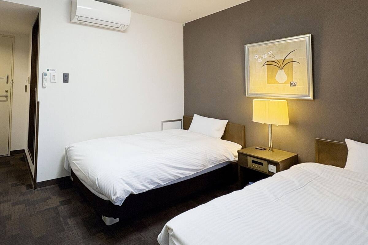 A hotel room with two beds, a bedside table with a lamp, an air conditioning unit, and a door leading to another room.