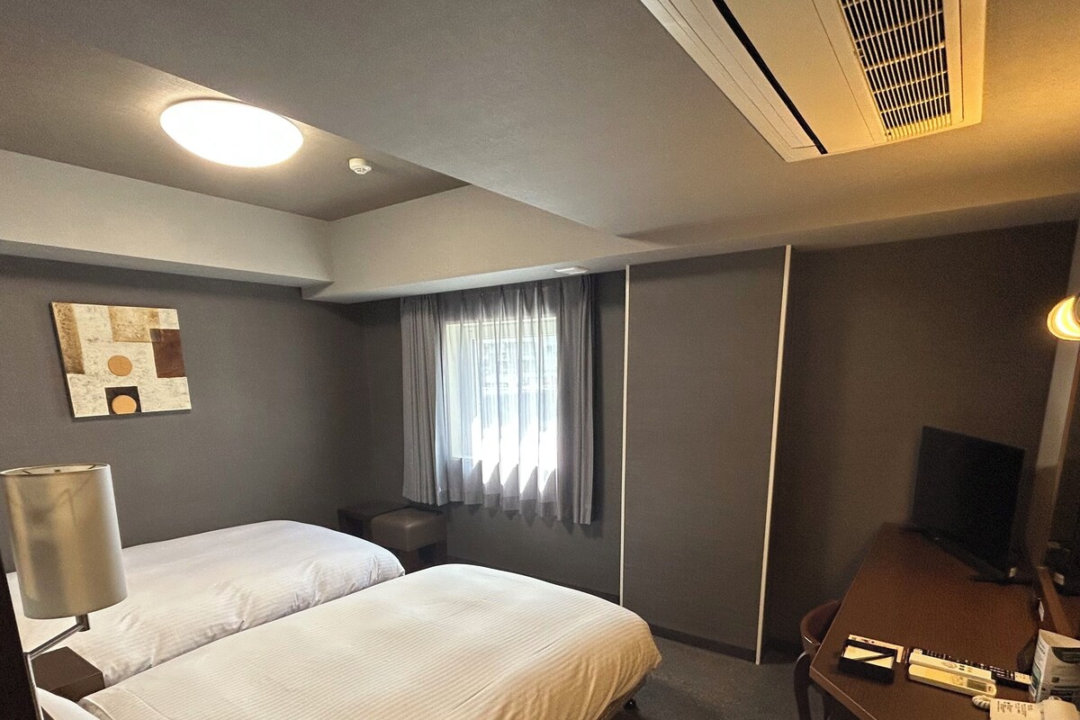 A hotel room with two beds, a desk, a chair, a lamp, and a window with curtains.