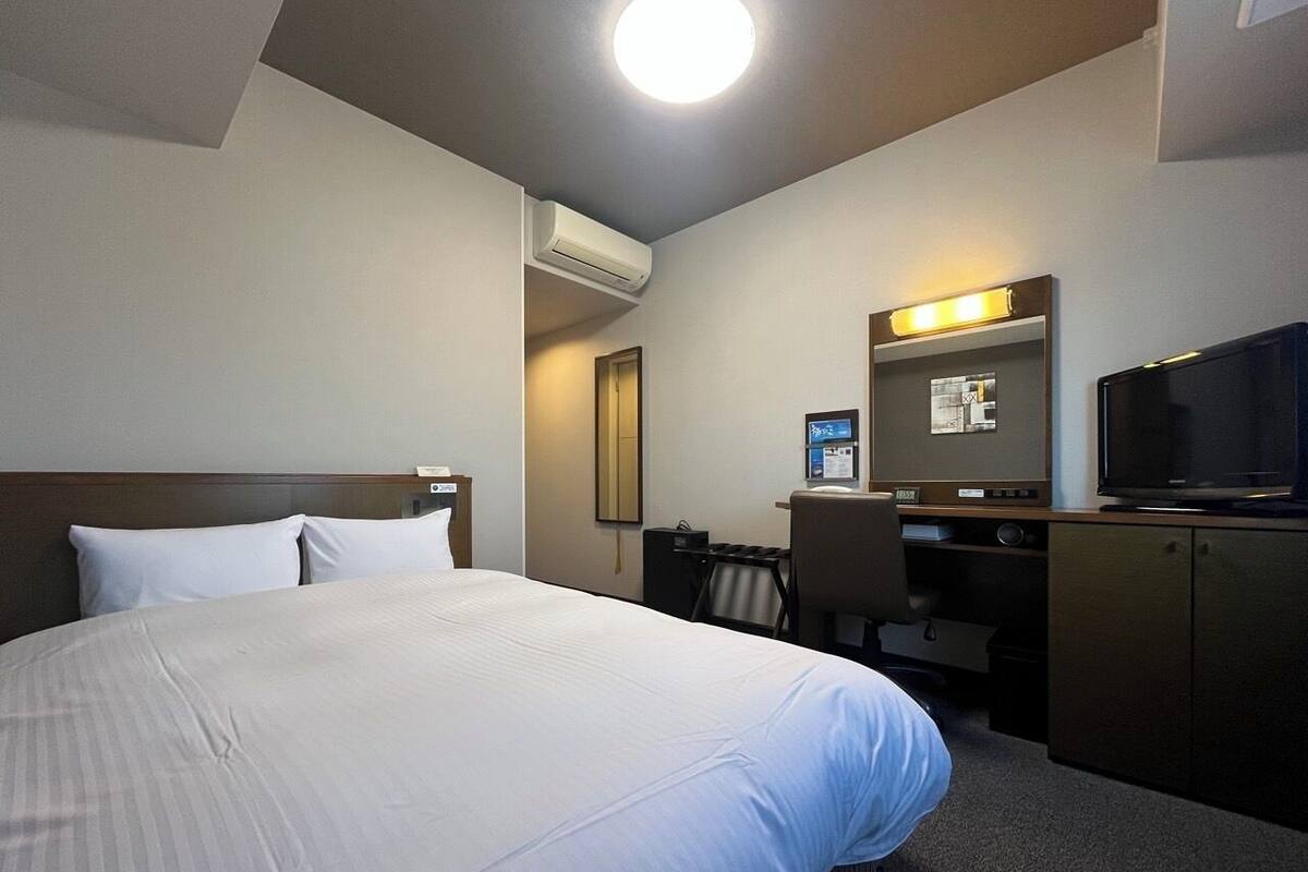 A hotel room with a bed, desk, chair, television, and an air conditioning unit.