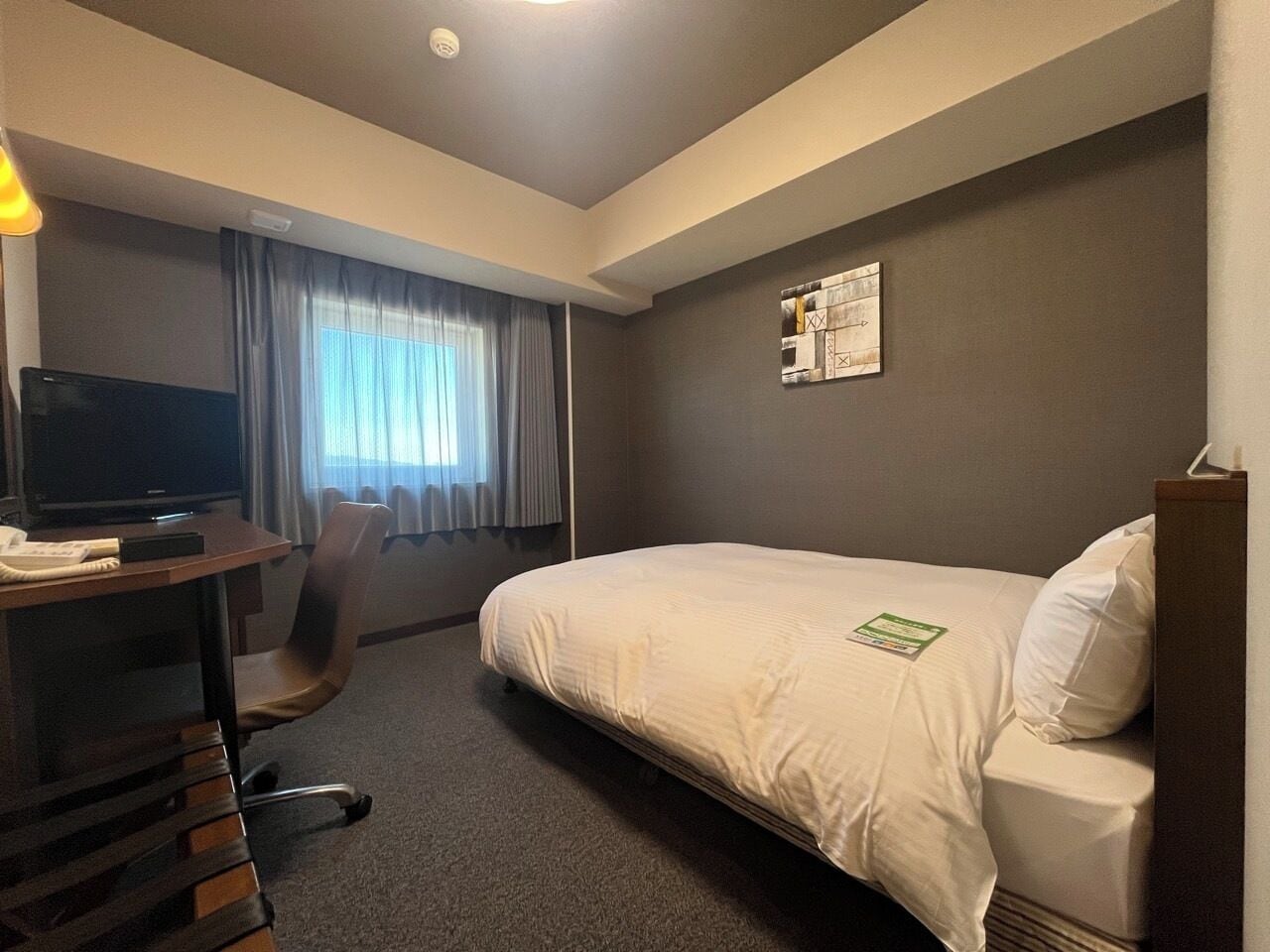 Ein Hotelzimmer mit einem Bett, einem Schreibtisch mit Stuhl, einem Fernseher und einem Fenster mit Vorhängen.