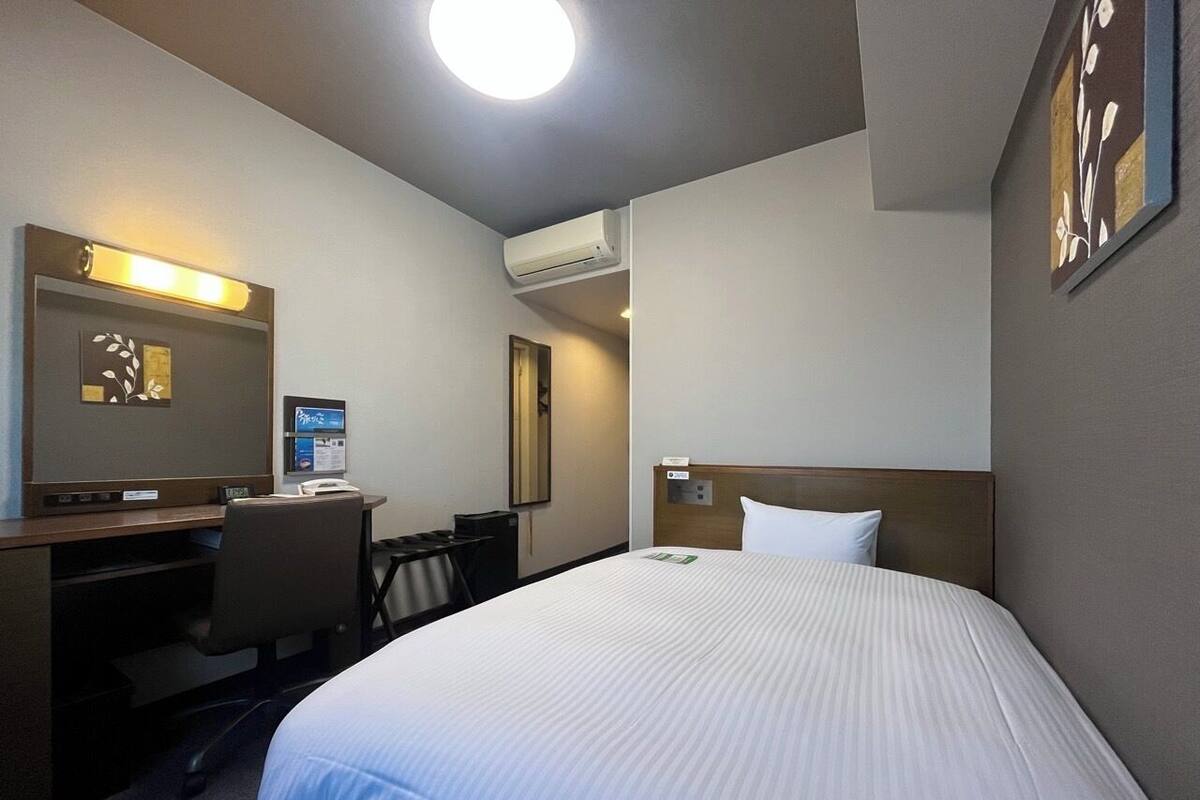 A hotel room with a bed, desk, chair, and a wall-mounted air conditioner.