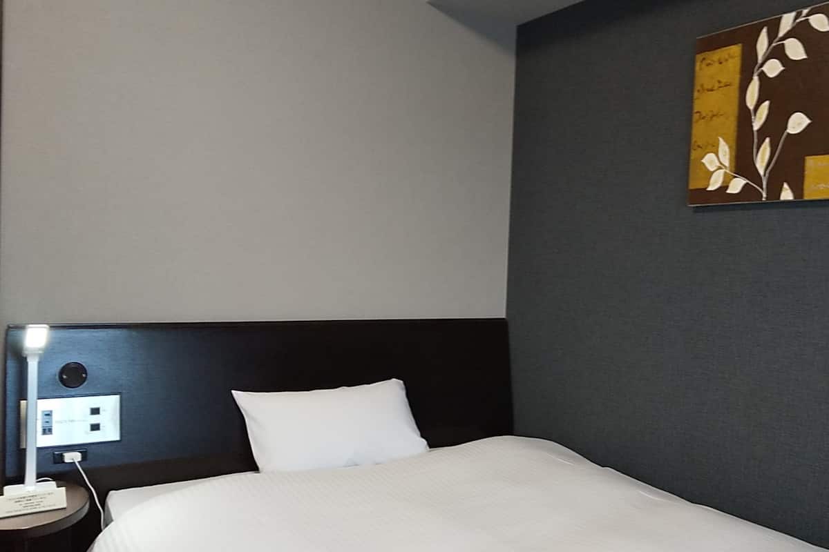 A hotel room with a bed, a nightstand with a lamp, a power outlet, and a wall with a decorative panel.