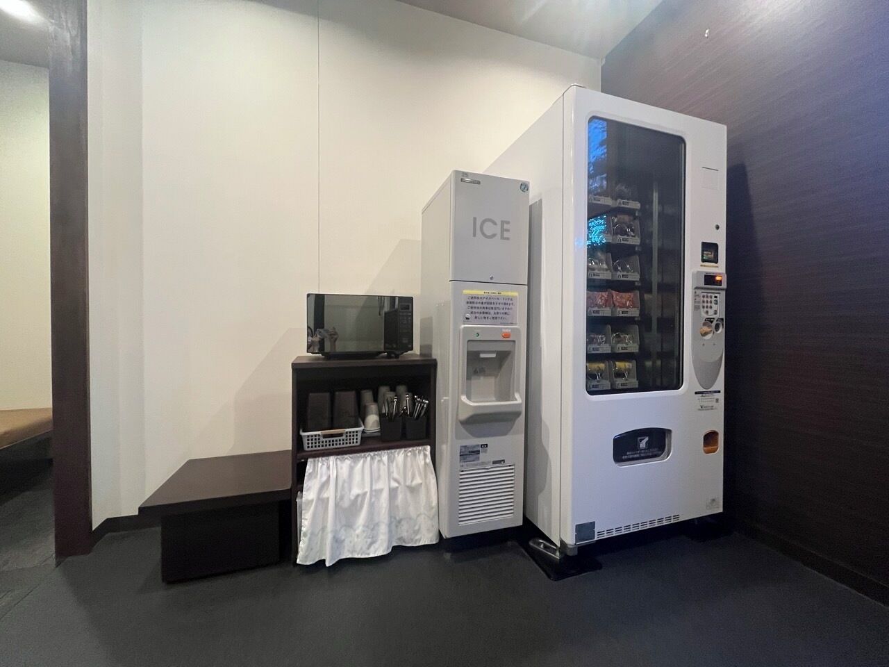 Una máquina expendedora con la etiqueta 'ICE' y un pequeño estante con vasos y utensilios.