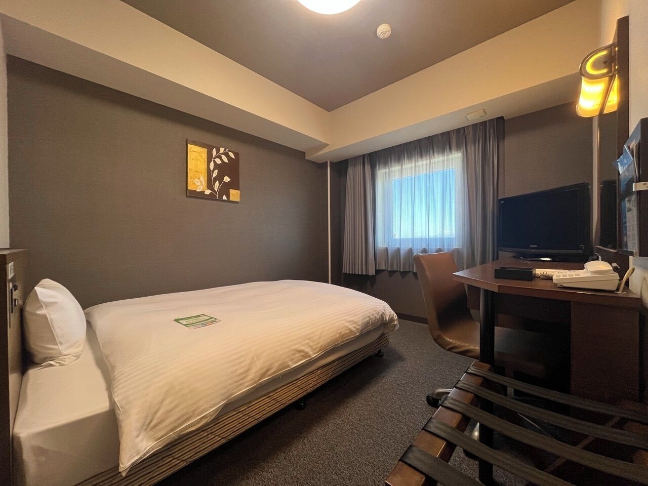 Una habitación de hotel con cama, escritorio con televisor, una silla y ventana con cortinas.