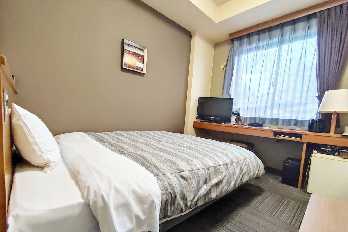 A hotel room with a bed, a desk with a laptop, a TV, and a window with curtains.