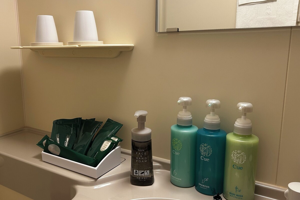 A bathroom with a toilet, a shelf with cups, and a row of toiletries including a hand soap dispenser, conditioner, and shampoo.