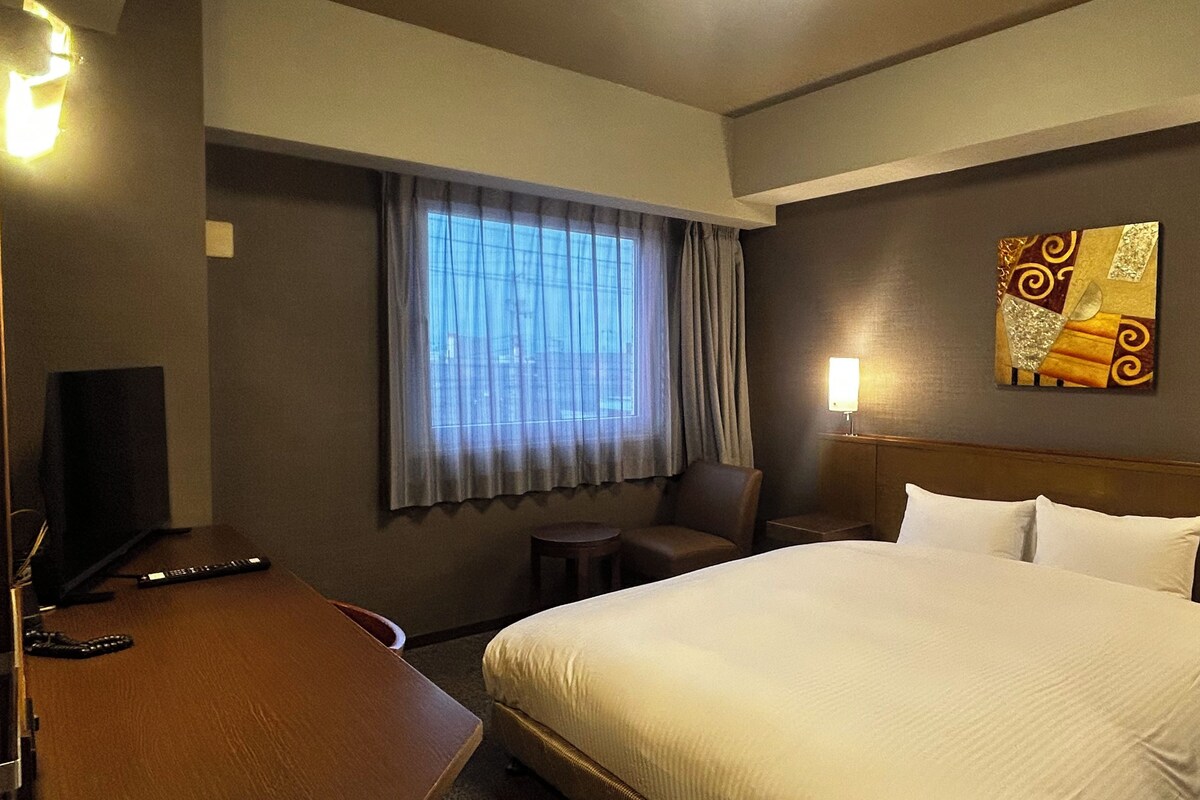 A hotel room with a large bed, a desk, a chair, a TV, a window with curtains, and a wall-mounted artwork.