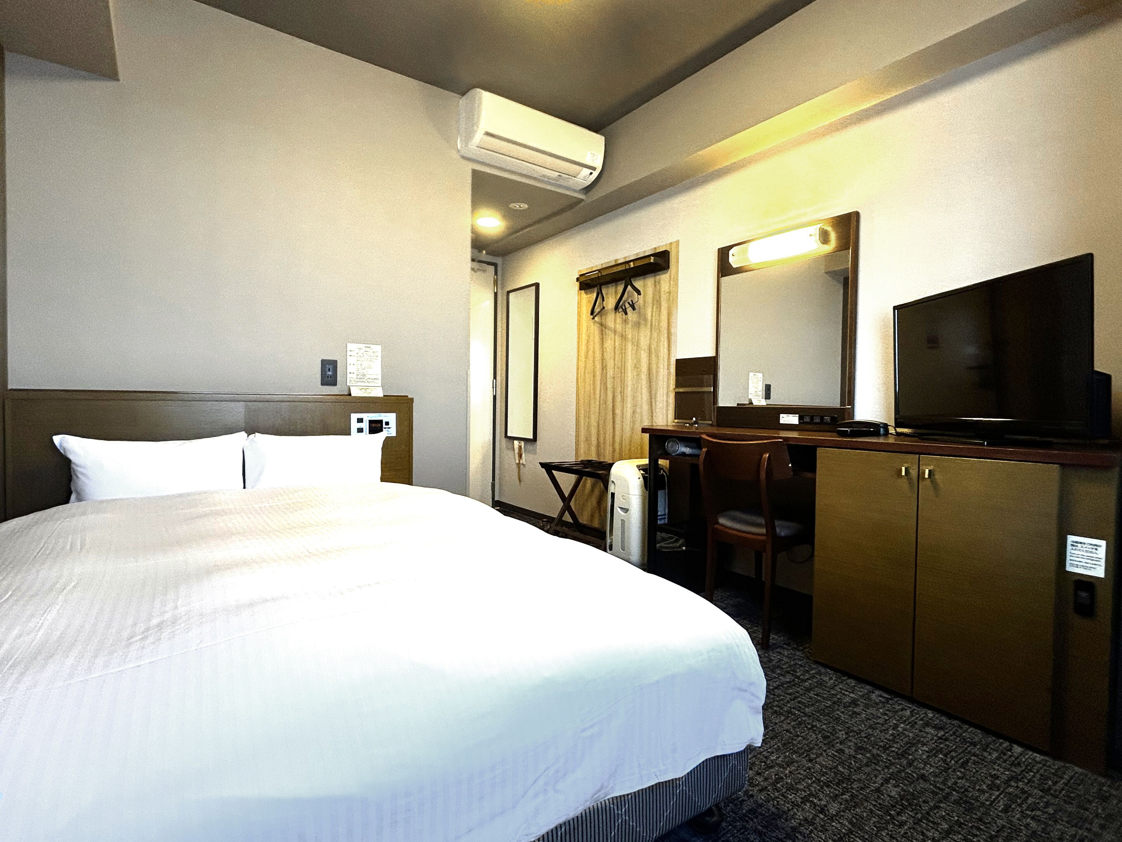 A hotel room with a bed, a desk, a chair, a television, and an air conditioning unit.