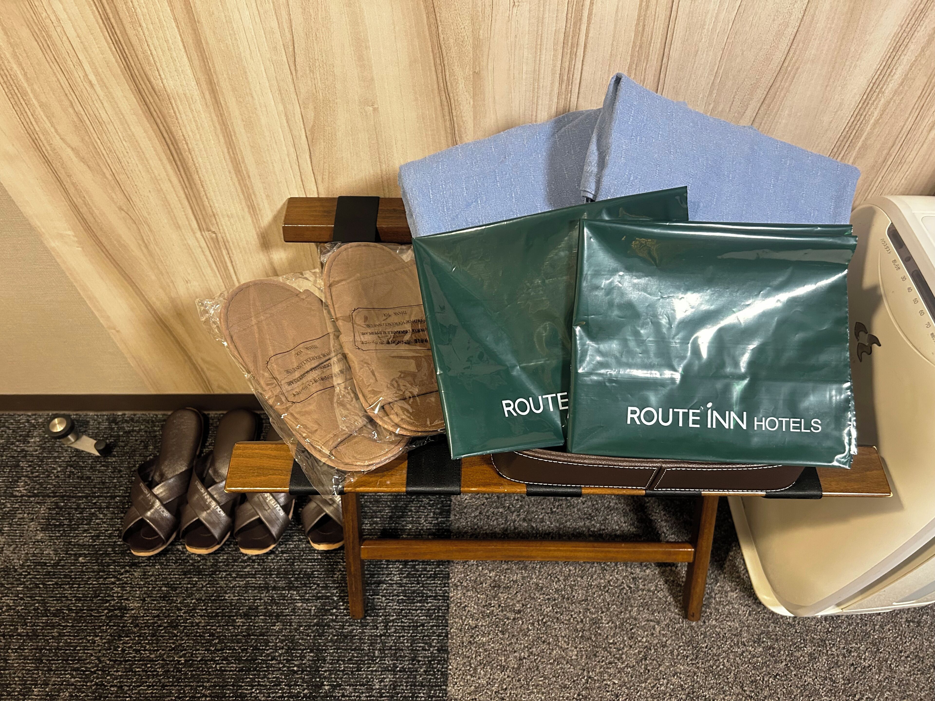 A room with a wooden floor, a pair of brown sandals, a black object, and a green bag with 'ROUTEINN HOTELS' text.