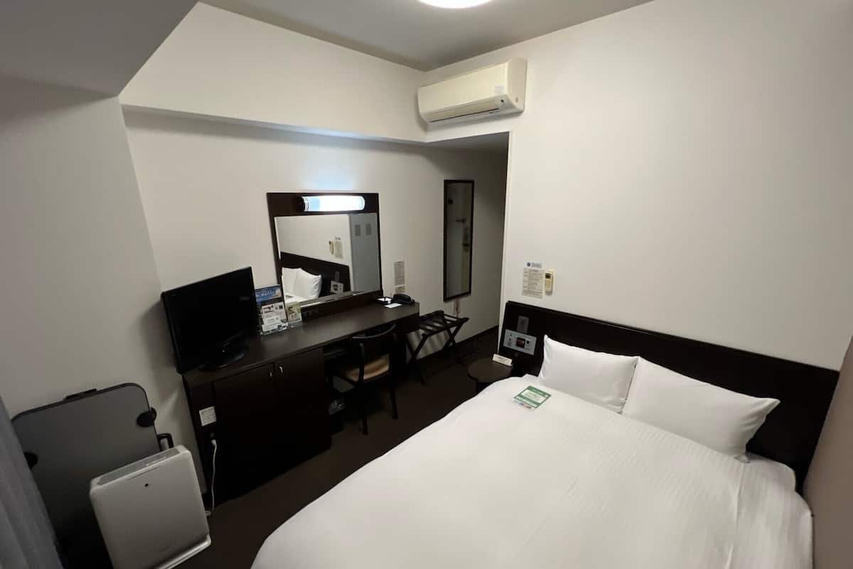 Ein Hotelzimmer mit einem Bett, einem Schreibtisch mit Computer, einem Stuhl, einem kleinen Tisch und einer Klimaanlage.