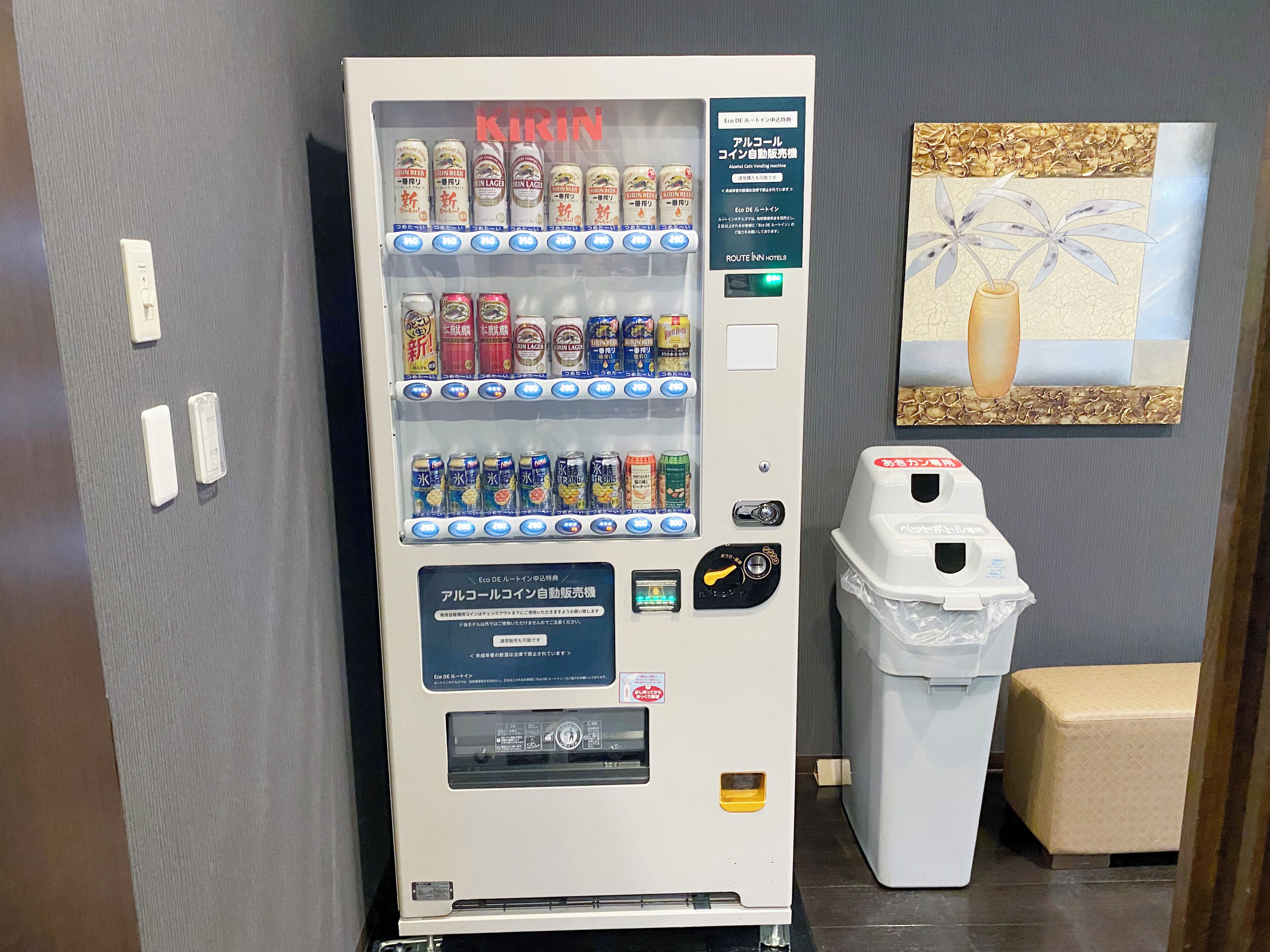 A vending machine with various drinks and snacks, a trash bin, and a painting on the wall.