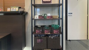 A hotel reception area with a reception desk, a sign indicating an amenity bar, and shelves with labeled baskets containing various items.