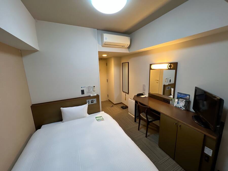 A hotel room with a bed, desk, chair, TV, and an air conditioning unit.