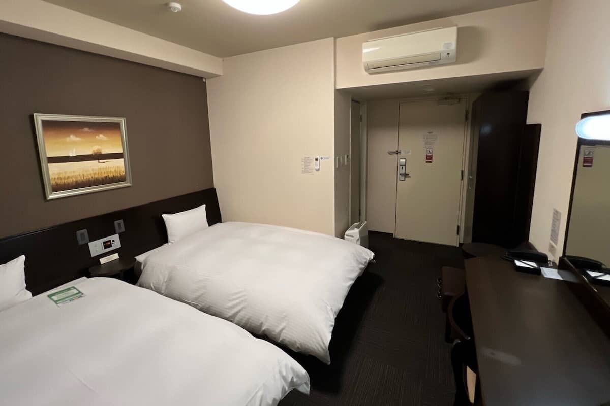 Ein Hotelzimmer mit zwei Betten, einem Schreibtisch, einem Spiegel, einer Klimaanlage und einer Tür zu einem weiteren Raum.