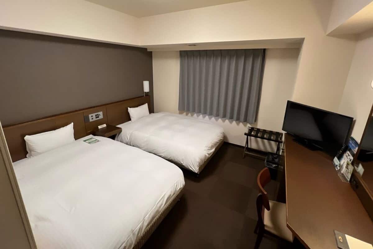 Ein Hotelzimmer mit zwei Betten, einem Schreibtisch, einem Fernseher und einem Vorhangfenster.