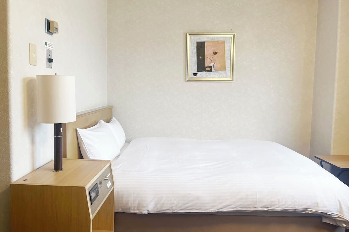 A hotel room with a bed, a bedside table with a lamp, a wall-mounted clock, and a framed picture on the wall.