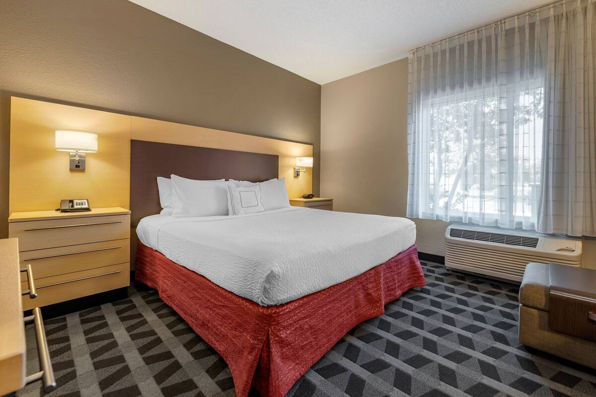 A hotel room with a large bed, two bedside tables with lamps, a desk, and a window with curtains.