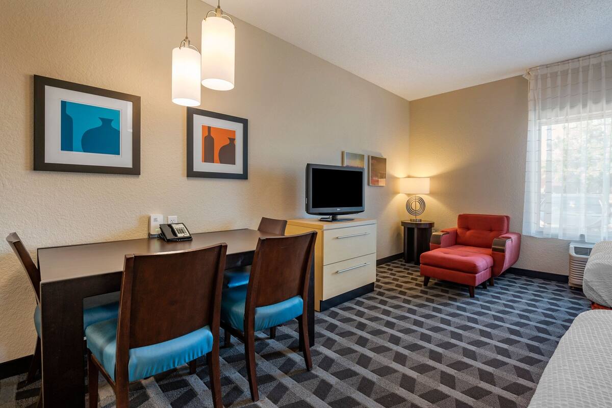 A hotel room with a desk, two chairs, a television, a red armchair, and artwork on the wall.