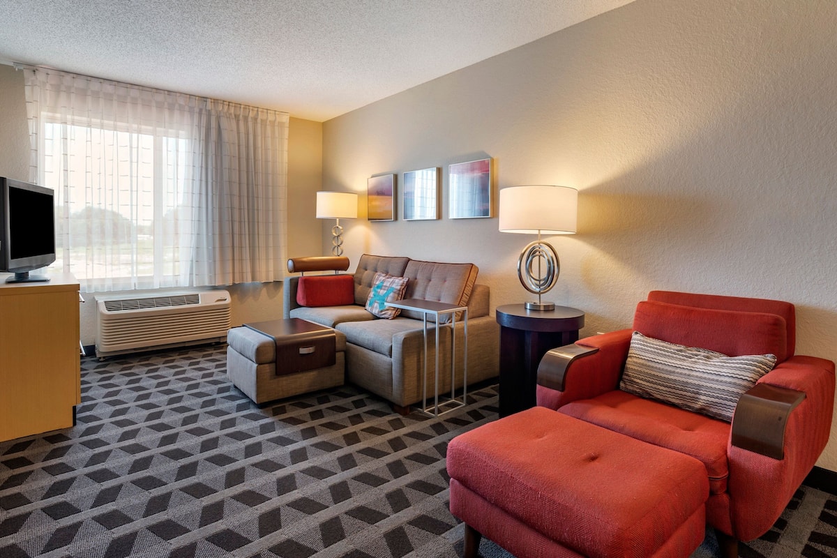 A hotel room with a sofa, armchair, TV, air conditioning unit, and artwork on the wall.