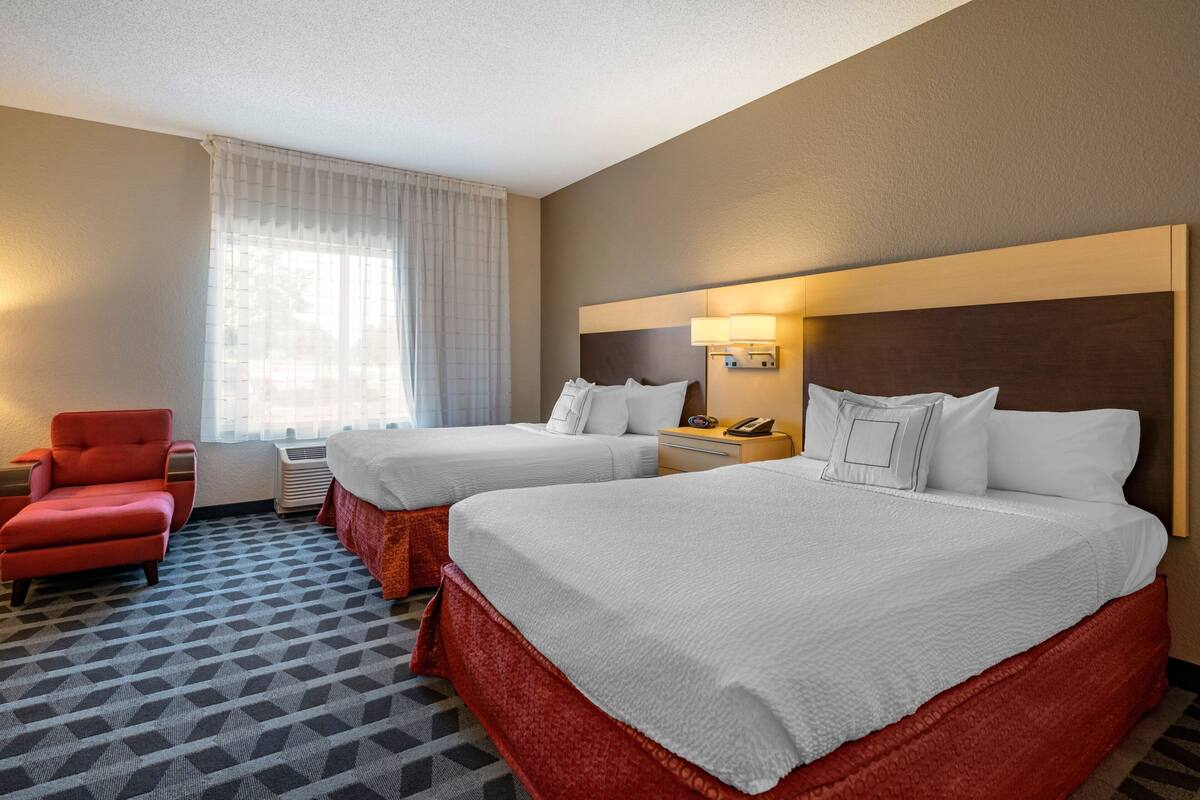 A hotel room with two beds, a red armchair, a bedside table with a lamp, and a window with curtains.
