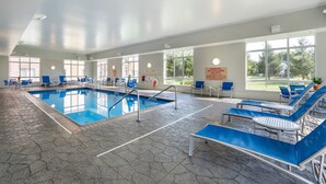 Una piscina interior con agua azul, rodeada de sillas y mesas de descanso azules, y ventanas amplias que dejan entrar luz natural.