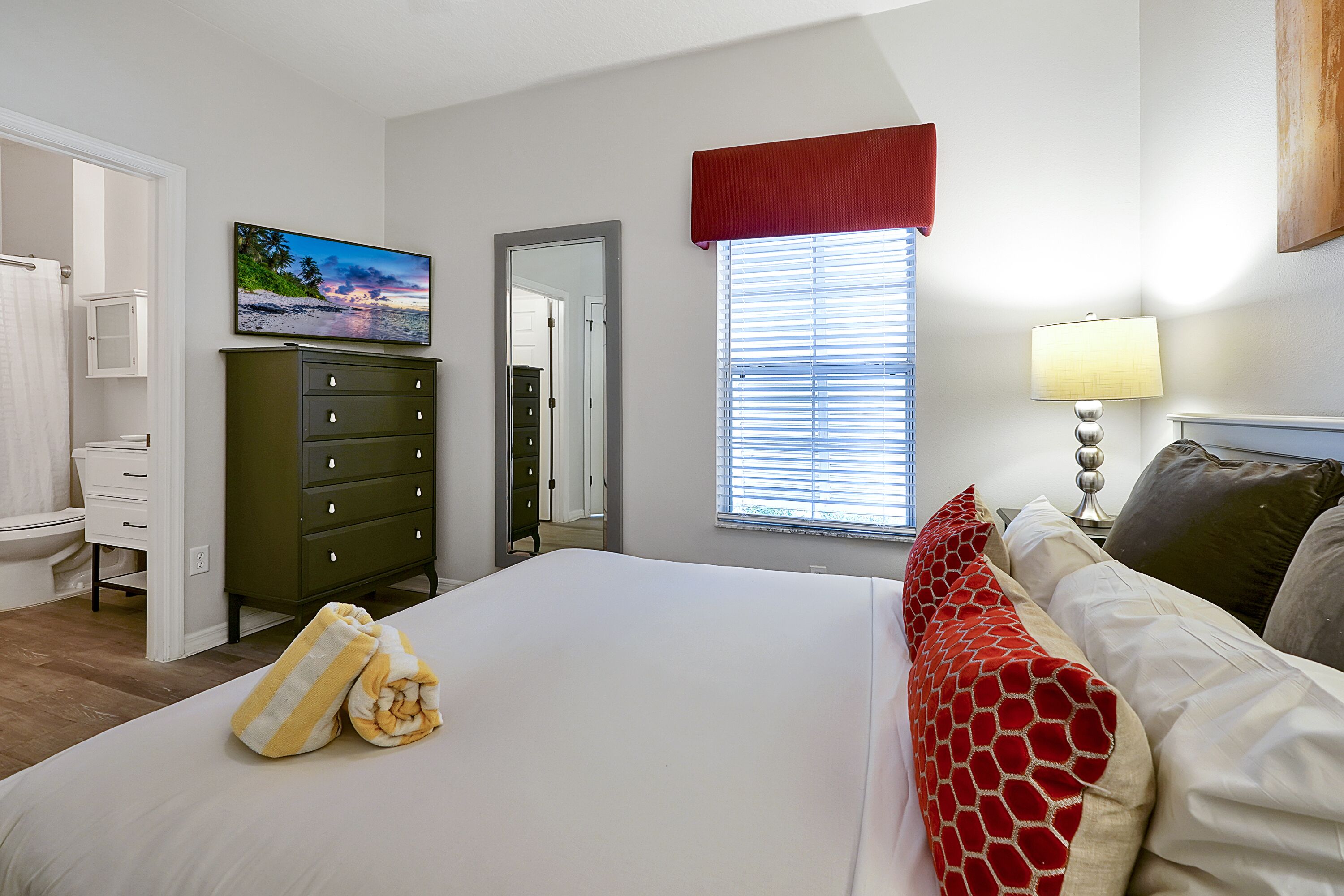 A bedroom with a bed, a dresser, a television, a window with blinds, and a lamp.