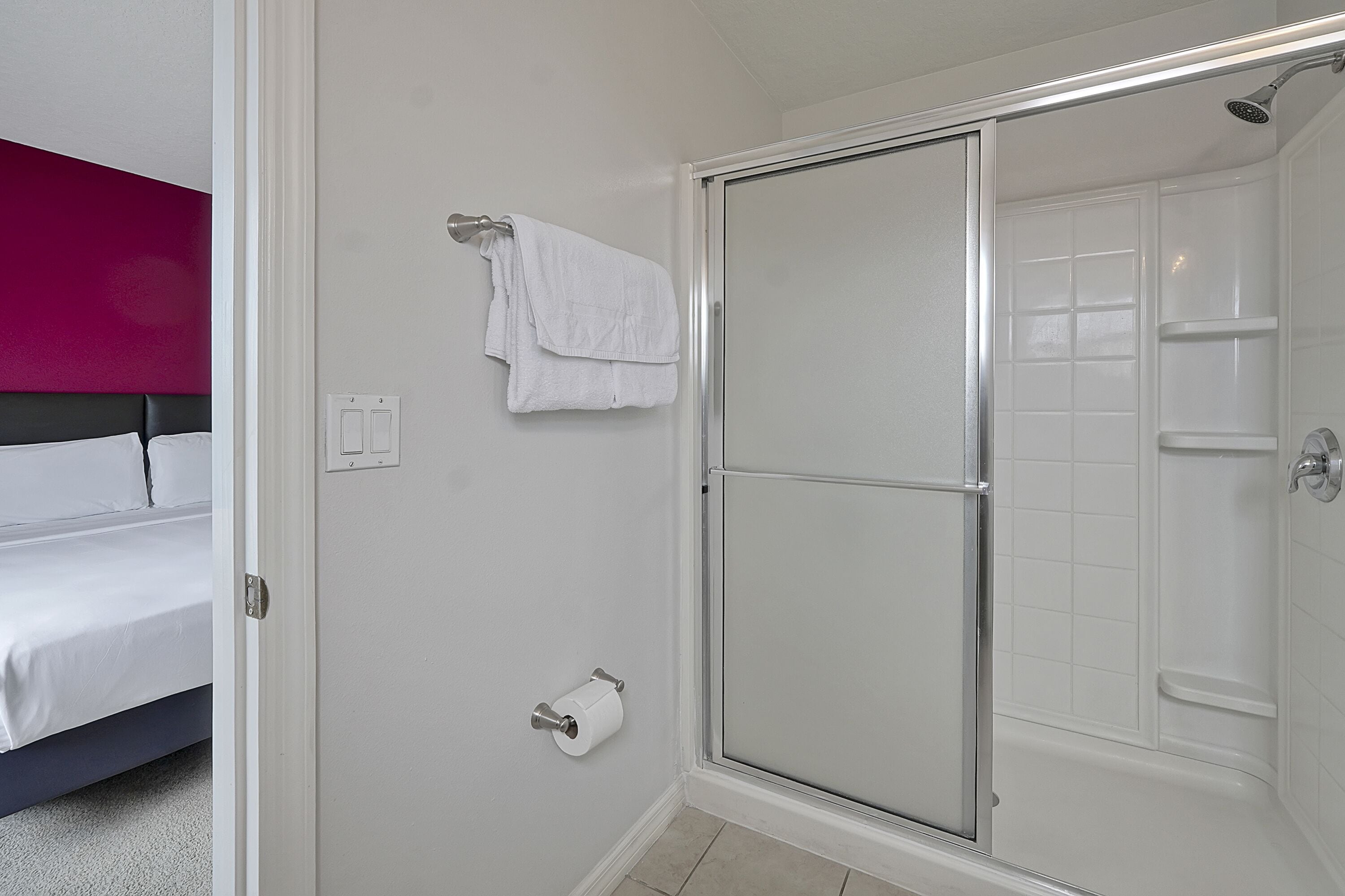 A hotel bathroom with a shower, toilet, and a bed with a headboard.