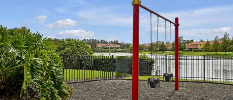 Un parque infantil con un columpio rojo y una cerca negra, rodeado de vegetación y un cuerpo de agua al fondo.