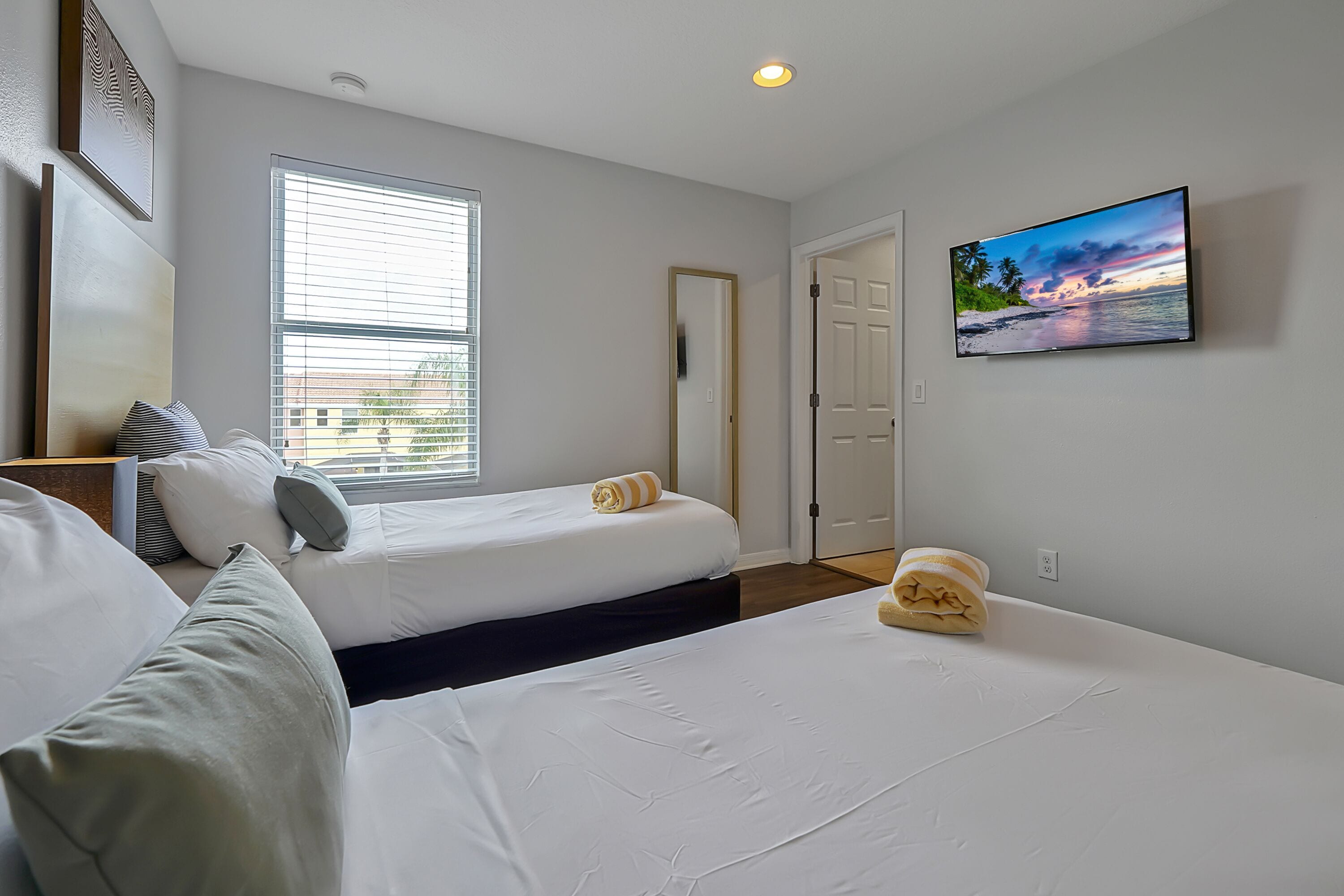 Habitación de hotel con dos camas, un ventanal con persianas, un televisor de pantalla plana en la pared y una puerta que da a otra habitación.