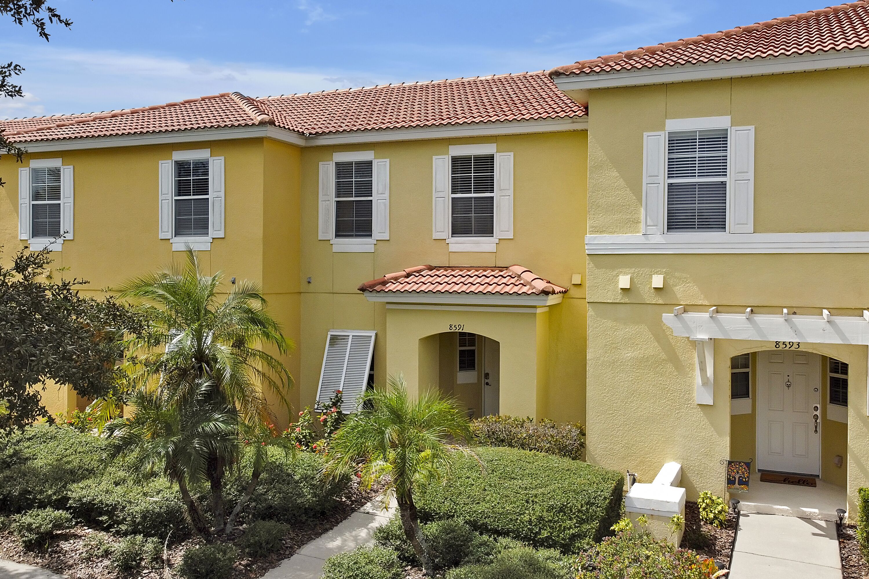 A yellow building with a red-tiled roof, white shutters, and a small garden with palm trees and shrubs.