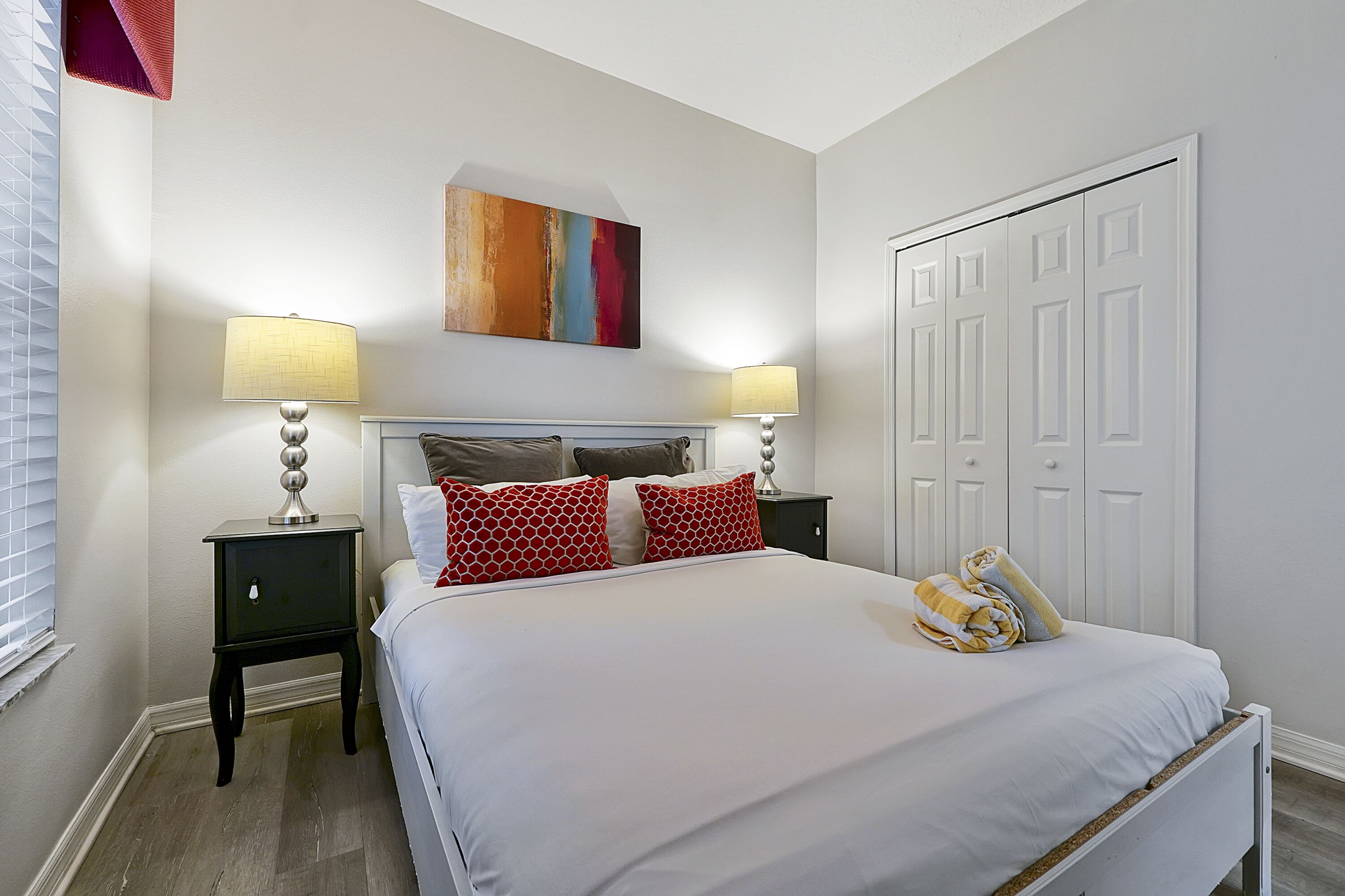 A bedroom with a large bed, two bedside tables with lamps, a colorful abstract painting above the bed, and a white door.