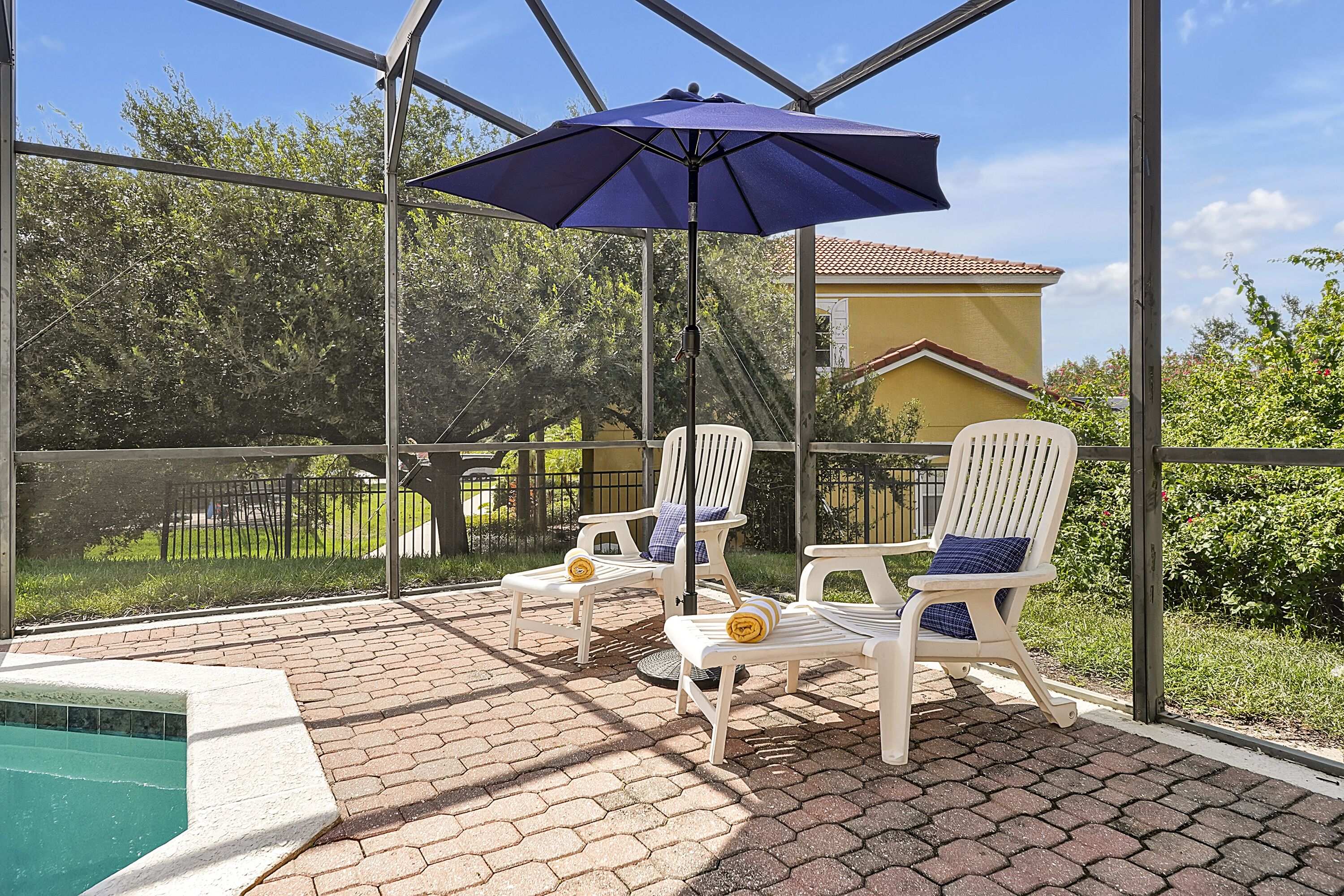 A covered outdoor area with a pool, two white chairs with blue cushions, and a blue umbrella.