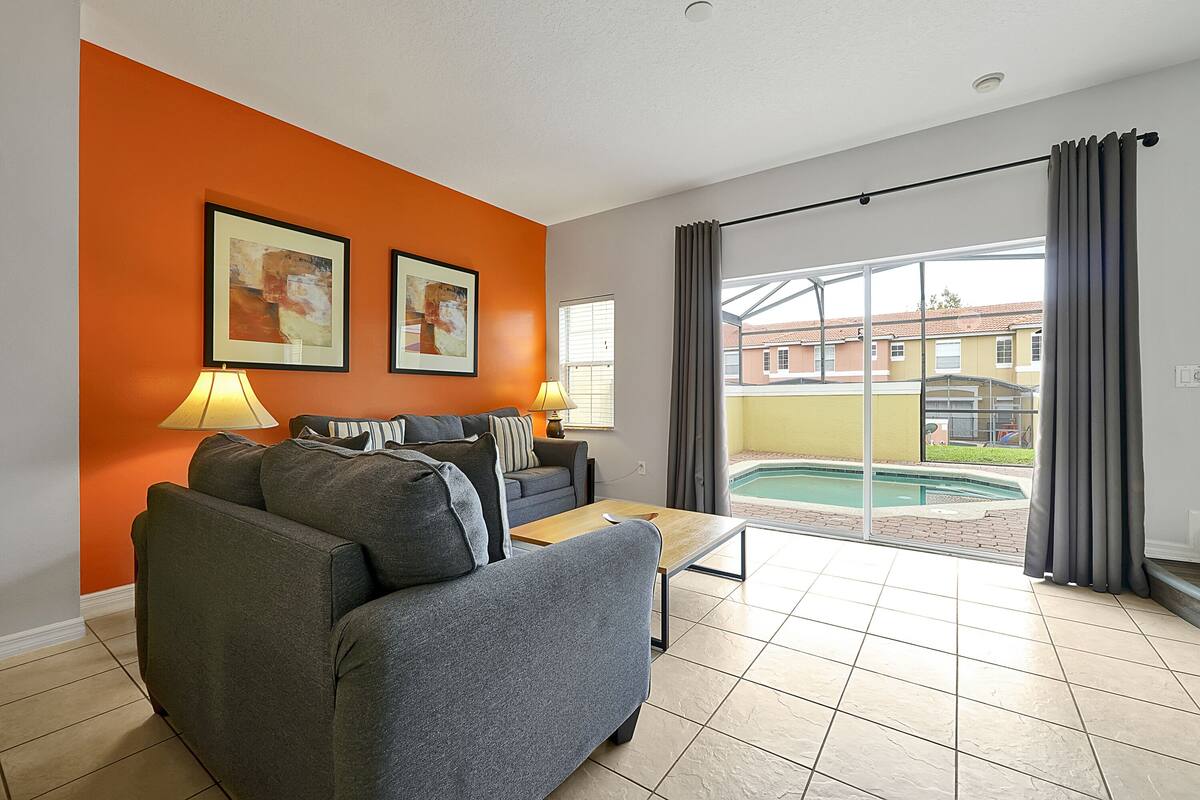 Ein Wohnzimmer mit grauem Sofa, einem Couchtisch und zwei Lampen. An der orangefarbenen Wand hängen zwei gerahmte Bilder. Eine Glastür führt zu einem Poolbereich.